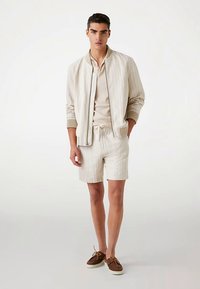 Lysefarvet bomberjakke med lodrette striber, kombineret med matchende shorts og en beige T-shirt med rund hals. Brune snørestøvler fuldender looket.