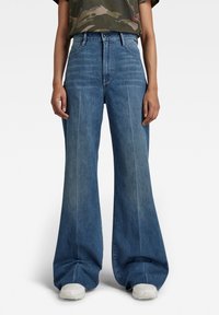 Široké modré džíny s vysokým pasem, které mají přední švy a jemnou texturu. Stylizované s tričkem s kamuflážním vzorem a teniskami.