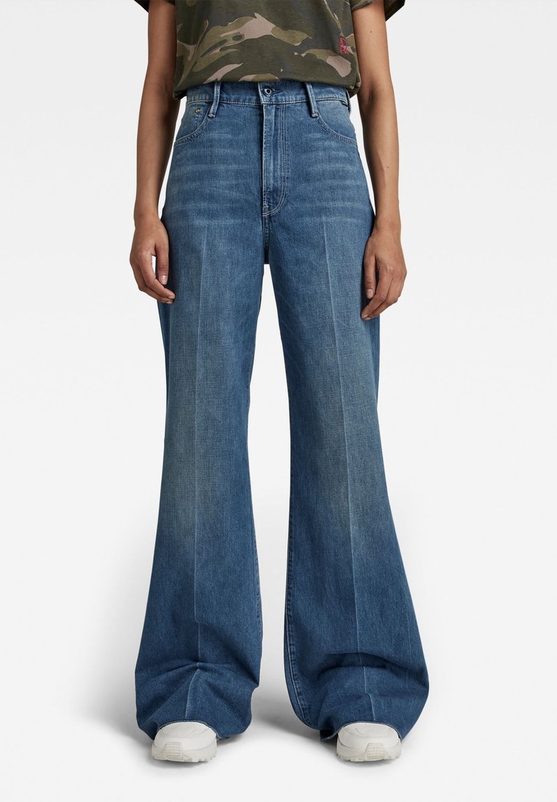 Široké modré džíny s vysokým pasem, které mají přední švy a jemnou texturu. Stylizované s tričkem s kamuflážním vzorem a teniskami.