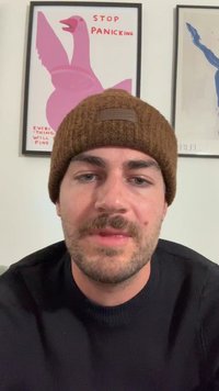 Szakállas férfi barna kötött sapkában és fekete pulóverben ül egy fedett térben, mögötte keretezett művészet látható, beleértve egy rózsaszín madarat és a "NE PÁNIKOLJ" feliratot.