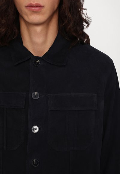 Veste en daim bleu foncé avec un col, fermeture à boutons sur le devant, deux poches poitrine et une texture lisse et mate.