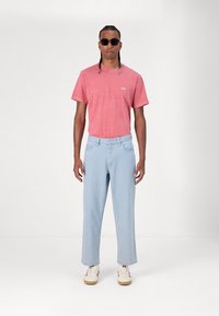 Ροζ κοντομάνικο μπλουζάκι με το λογότυπο "obey", συνδυασμένο με ανοιχτό μπλε φαρδύ παντελόνι τζιν και λευκά αθλητικά παπούτσια με κόκκινες ρίγες.