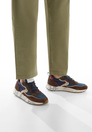 Personne portant un pantalon olive et des baskets bleu marine et marron avec des semelles blanches, ornées de texte en gras sur le côté.