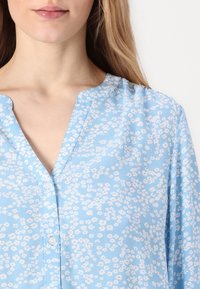 Blusa azzurra chiara con motivo floreale, scollatura a V e bottoni frontali. Realizzata in tessuto morbido con una leggera lucentezza.