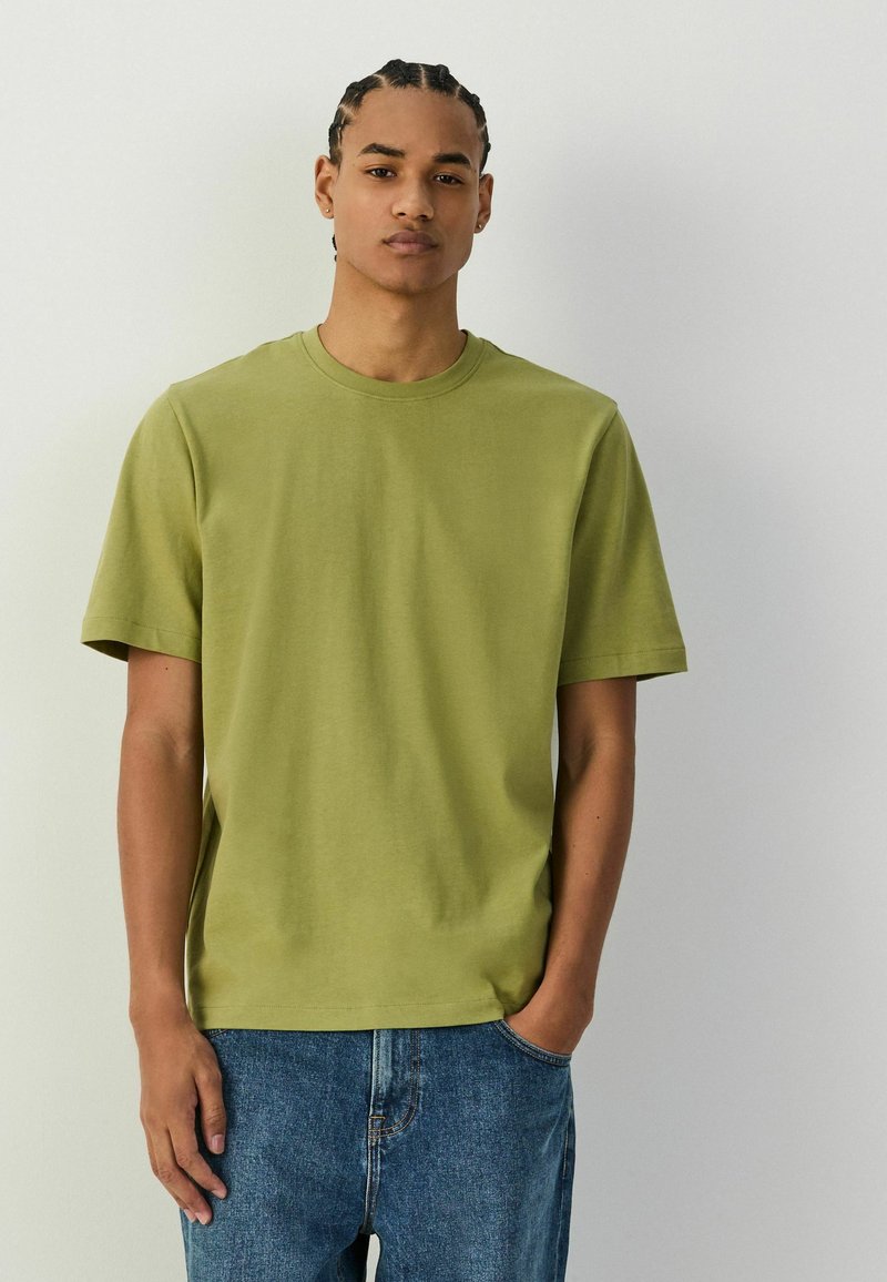 Jeune homme aux cheveux tressés portant un T-shirt manches courtes vert olive et un jean bleu, debout devant un fond clair uni.