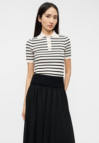 Ριγέ πόλο μπλουζάκι με κοντά μανίκια σε μαύρο και κρεμ χρώμα, με κλασικό γιακά και τρία κουμπιά, συνδυασμένο με μαύρη φούστα ψηλής μέσης.