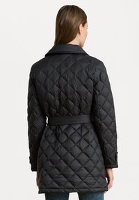 Zwarte gewatteerde pufferjas met een ceintuur in de taille, grote kraag, en knoopaccenten op de mouwen, met ruitvormige stiksel.