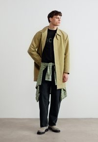 Άνδρας που στέκεται φορώντας καμηλό καμπαρντίνα πάνω από μαύρο πουκάμισο, με πράσινο καρό πουκάμισο δεμένο στη μέση, μαύρο παντελόνι και μπεζ παπούτσια, μπροστά σε λευκό τοίχο.