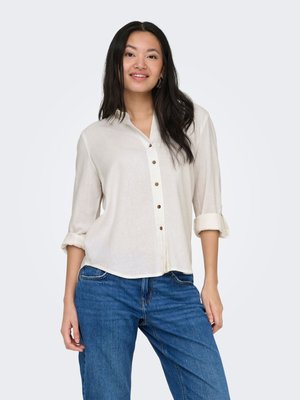 Femme aux longs cheveux foncés portant une chemise blanche boutonnée avec manches retroussées et un jean bleu, souriante devant un fond clair uni.