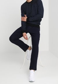 Marinblå texturerad tröja med svart ribbad fåll, kombinerad med marinblå byxor och vita sneakers, sittande på en vit stol mot en ljus bakgrund.