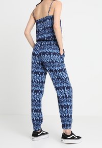 Niebieski kombinezon we wzory z cienkimi ramiączkami, elastycznym pasem i kieszeniami; zwężane nogawki i zakończone mankietami; w zestawie z czarnymi sneakersami.