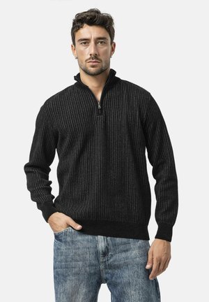 Black ribbed zip-up sweater with a high collar, made from a textured knit fabric. Paired with light blue denim jeans.