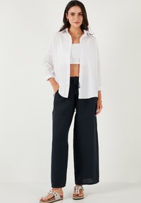 Camicia bianca a bottoni, pantaloni neri in lino larghi e sandali con motivo. L'outfit ha una vestibilità rilassata e un design minimalista.