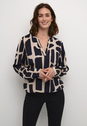 Blusa de mangas largas y escote en V en beige con un patrón geométrico abstracto en azul marino. Cuenta con mangas fruncidas y puños abotonados.