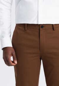 Braune Chinos aus strukturiertem Stoff mit geradem Schnitt, ausgestattet mit einem Frontknopfverschluss und Seitentaschen. Sichtbares Bündchen eines weißen Hemdes.