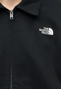 Veste noire zippée avec col, arborant un logo "The North Face" brodé en blanc sur la poitrine gauche et une tirette de fermeture assortie.