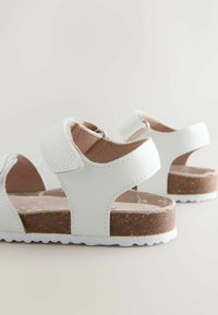 Sandale albe din piele, cu bretele reglabile, talpă interioară din plută și talpă exterioară din cauciuc alb. Prezintă un design decupat și căptușeală interioară moale.