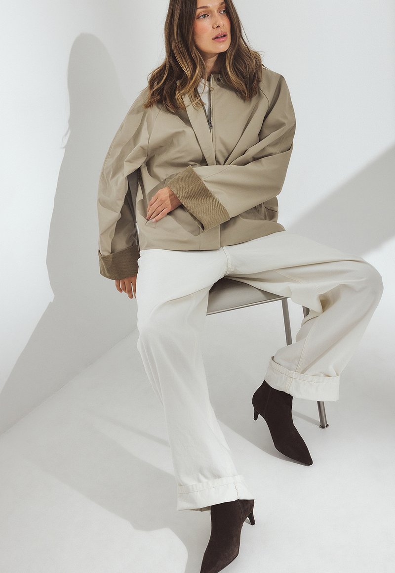 Chaqueta oversize color caqui con mangas anchas, combinada con pantalones blancos. Presenta botas de tacón puntiagudas en marrón oscuro. Texturas suaves, diseño minimalista.