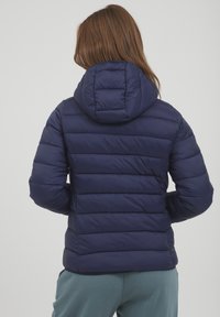 Námornícka puffer bundu s horizontálnym švom a kapucňou. Hladký povrch s lesklým vzhľadom, obsahuje nastaviteľnú kapucňu pre väčšie teplo.