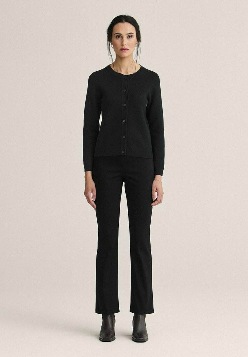 Svart stickad cardigan med rund halsringning och knäppning, i kombination med svarta åtsittande byxor och ankelboots. Enkel, strömlinjeformad design.