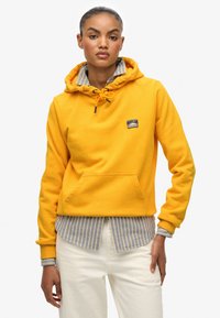 Felpa di cotone gialla con coulisse e tasca frontale, indossata sopra una camicia a righe blu e bianche, abbinata a pantaloni bianchi.