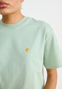 T-shirt di cotone verde chiaro con collo a giro e maniche corte. Logo giallo ricamato sul petto. Texture liscia.
