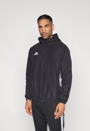 Czarna kurtka zapinana na zamek z kapturem, wykonana z gładkiej tkaniny. Na bokach znajdują się białe logo Adidas i paski, a mankiety są elastyczne.