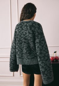 Sivi, veliki pulover s pahuljastom teksturom, koji ima uzorak leoparda i suptilne sjaje. S rebrastim donjim rubom i dugim rukavima.