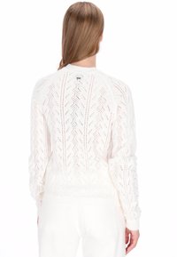 Pull en tricot blanc avec un motif texturé et ajouré, col rond et poignets côtelés. Le dos présente un petit logo.