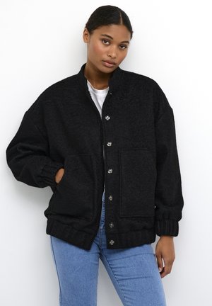 Veste noire surdimensionnée avec une finition texturée, boutons-pression, manches élastiques et deux poches avant. Portée par-dessus un haut blanc et un jean bleu.