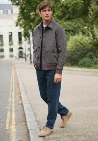 Young man in a brown checkered jacket, navy shirt, blue jeans, and beige shoes walking on a city sidewalk near greenery and buildings.