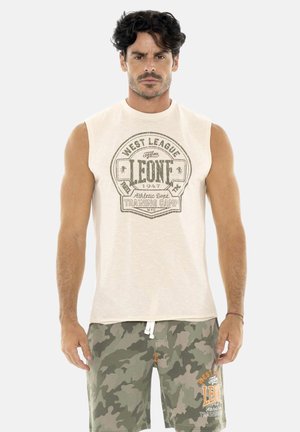 Homme aux cheveux bouclés foncés portant un débardeur beige avec l'impression "LEONE Training Camp" et un short camouflage vert avec logo assorti.