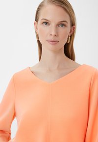 V-hals, langemouwtop in fel oranje stof met een gladde textuur en een centrale verticale naad. Gouden oorbellen met een textuur in de vorm van een ring.
