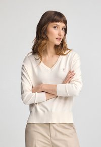 Maglione cream a V con maniche e orlo a coste. Logo ricamato discreto sul petto. Morbido al tatto, vestibilità comoda. Abbinato a pantaloni beige.