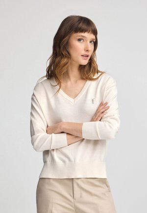 Jersey crema con escote en V y puños y dobladillo acanalados. Sutil logo bordado en el pecho. Textura suave, corte holgado. Combinado con pantalones beiges.
