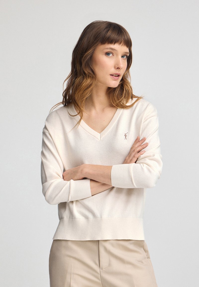 Maglione cream a V con maniche e orlo a coste. Logo ricamato discreto sul petto. Morbido al tatto, vestibilità comoda. Abbinato a pantaloni beige.