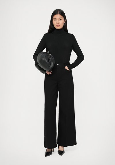 Kvinde iført en sort turtleneck, brede sorte bukser, sorte hæle, holder en stor sort håndtaske mod en ensfarvet baggrund.
