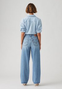 Femme aux cheveux courts et bruns, portant une chemise bleu clair retroussée, un jean taille haute à jambes larges, et des chaussures beiges à talons hauts, debout de dos.