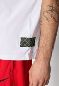 Close-up of a person’s arm with a star and barbed wire tattoo, wearing a white shirt with a black and green stitched patch, and red shorts.