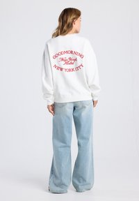 Sudadera blanca con texto bordado en rojo en la parte trasera que dice "GOODMORNING New York Hotel New York City", combinada con jeans de pierna ancha de color azul claro.