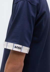 Tmavomodré polo tričko s krátkym rukávom má biely pruhovaný lem s modrým vyšitým logom. Vyrobené z jemnej bavlnenej tkaniny, má klasický dizajn.