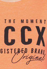 T-shirt arancione a maniche corte con testo nero in grassetto che recita "IL MOMENTO CCX MARCHIO REGISTRATO Originale." Tessuto morbido, design con collo a giro.