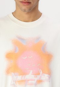 Šviesios spalvos medvilninė marškinė su pasteliniu saulės dizainu, kuriame yra rožinės ir oranžinės atspalvių, taip pat stilizuotas tekstas kontrastingos spalvos.
