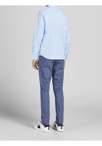 Světle modrá košile s dlouhým rukávem a zádovým dílem, kombinovaná s tmavě modrými úzkými kalhotami a bílými teniskami s texturovaným pruhovaným akcentem.