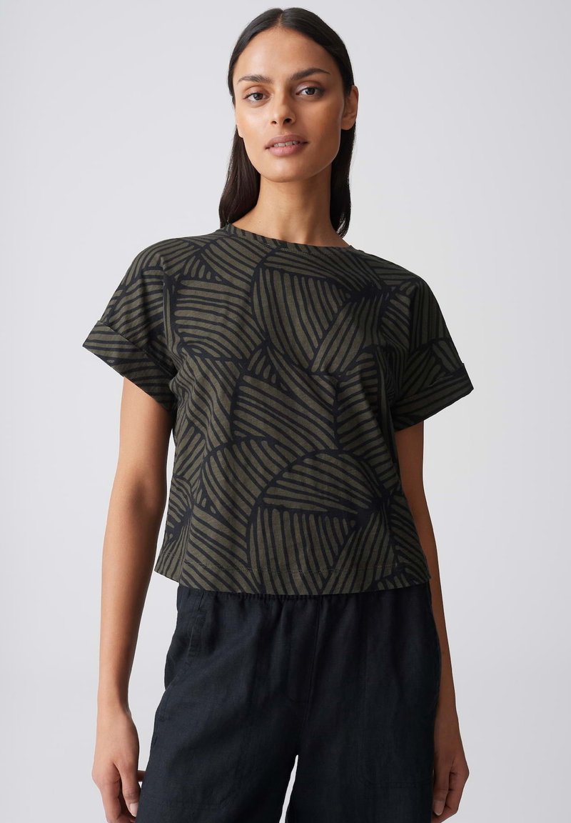 Frau trägt ein olivgrünes Kurzarmshirt mit schwarzen abstrakten geometrischen Mustern und lockere schwarze Hosen vor einem einfarbigen Hintergrund.
