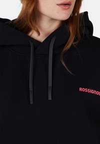 Felpa nera con cappuccio e cordoncino, caratterizzata da una texture liscia e un audace logo rosa "ROSSIGNOL" sul davanti. Inquadratura ravvicinata del collo.