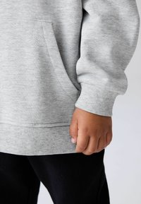 Felpa grigia con tasca frontale e polsini a coste, abbinata a pantaloni neri. Il tessuto è morbido e presenta un design classico e rilassato.