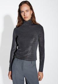 Maglietta nera a maniche lunghe con collo alto realizzata in un tessuto luccicante. Presenta una vestibilità slim e una texture liscia, abbinata a pantaloni grigi.