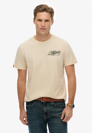 Camiseta de algodón color crema con mangas cortas, que presenta un gráfico de montaña verde oscuro y texto en el lado izquierdo del pecho. Llevada con vaqueros azules.