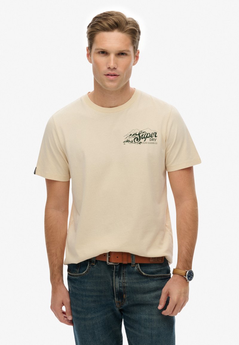 Camiseta de algodón color crema con mangas cortas, que presenta un gráfico de montaña verde oscuro y texto en el lado izquierdo del pecho. Llevada con vaqueros azules.